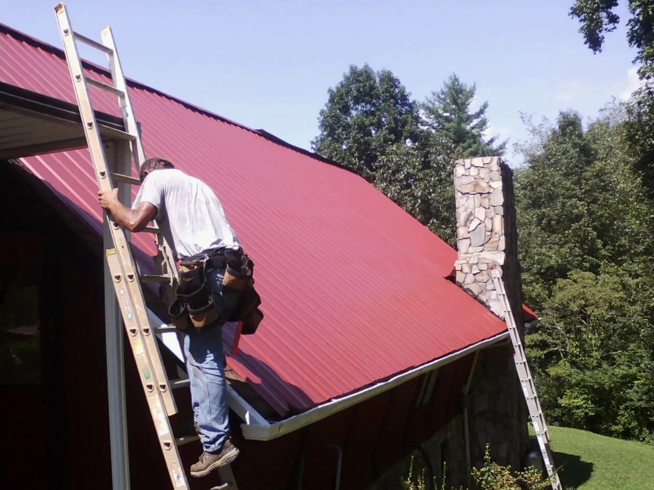 Roofer on a ladder beside a red metal roof and stone chimney in Mc Intosh
